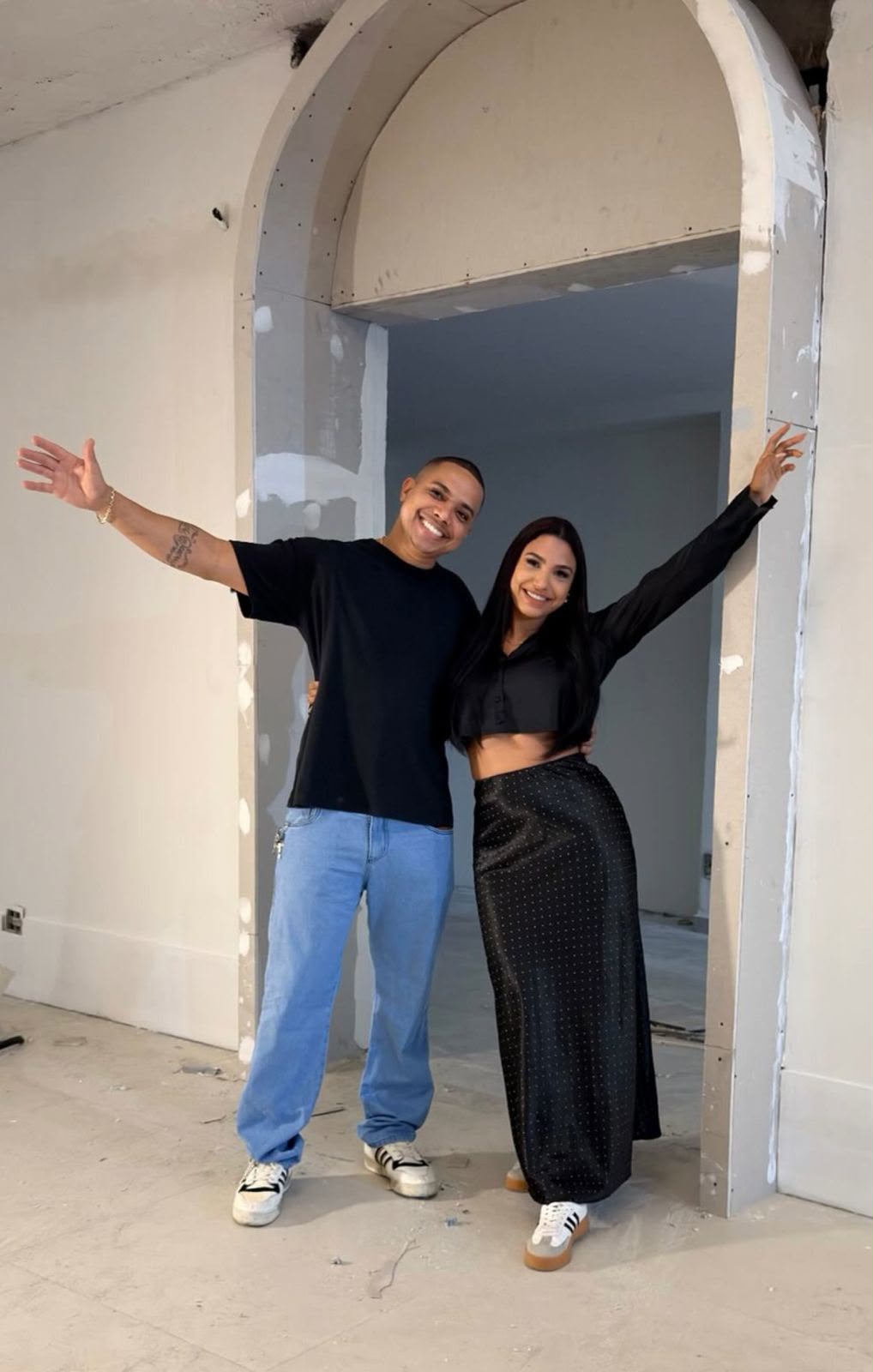 Tom e Raquel juntos, de braços abertos, em um espaço em construção